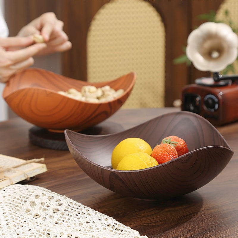 Two wooden bowl-shaped serving dishes on a dark wood table, with one in the foreground holding lemons and strawberries and the other in the background holding nuts. A person's hands are visible in the upper left holding food over the rear bowl.