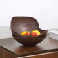 A dark brown wooden bowl with a wavy, layered design sits on a wooden table, containing lemons and strawberries. The background is a clean white wall with a book partially visible on the right.