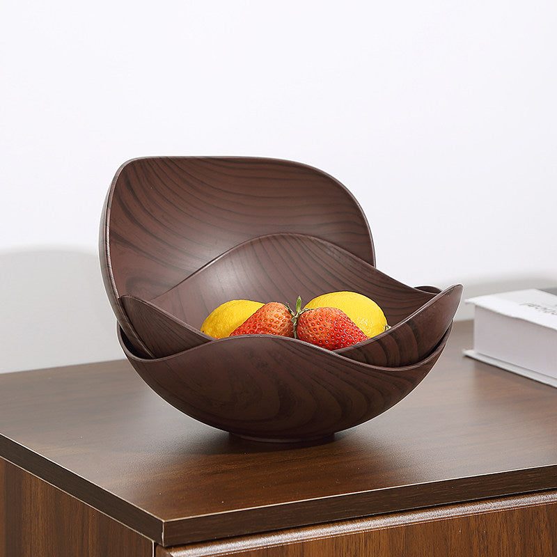 A dark brown wooden bowl with a wavy, layered design sits on a wooden table, containing lemons and strawberries. The background is a clean white wall with a book partially visible on the right.
