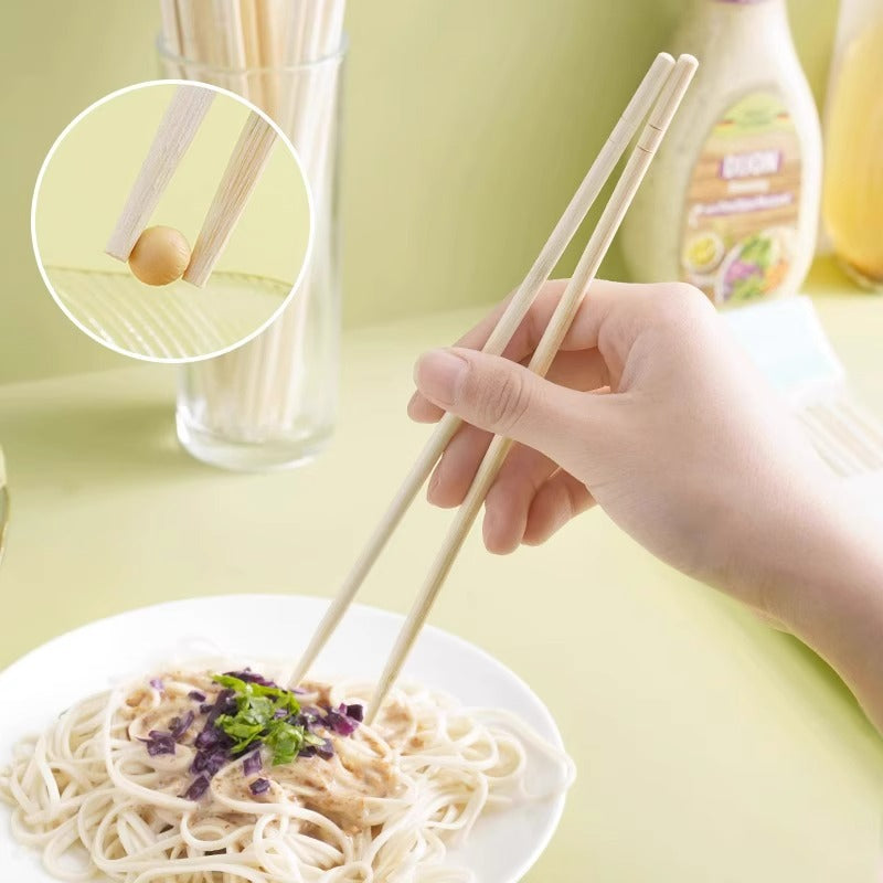 A hand holds a pair of light wood chopsticks over a plate of noodles, with a circular inset showing the chopsticks gripping a small food piece. The background is a soft pale green with a glass of chopsticks and a bottle blurred behind.