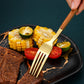 A close-up of a hand holding a gold-toned fork with a wooden handle, using it to lift a piece of steak from a black plate with gold veining. The plate is styled with grilled corn, tomato slices, and lime wedges on a dark textured background.