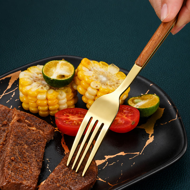 A close-up of a hand holding a gold-toned fork with a wooden handle, using it to lift a piece of steak from a black plate with gold veining. The plate is styled with grilled corn, tomato slices, and lime wedges on a dark textured background.