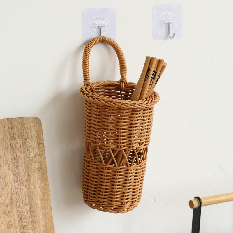 A cylindrical woven rattan pencil holder with a top handle, mounted on a white wall with two clear hooks. Wooden chopsticks are tucked inside the holder.