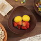A dark brown teardrop-shaped wooden bowl filled with two yellow lemons and several red strawberries, set on a wooden table with other food props and a book in the background.