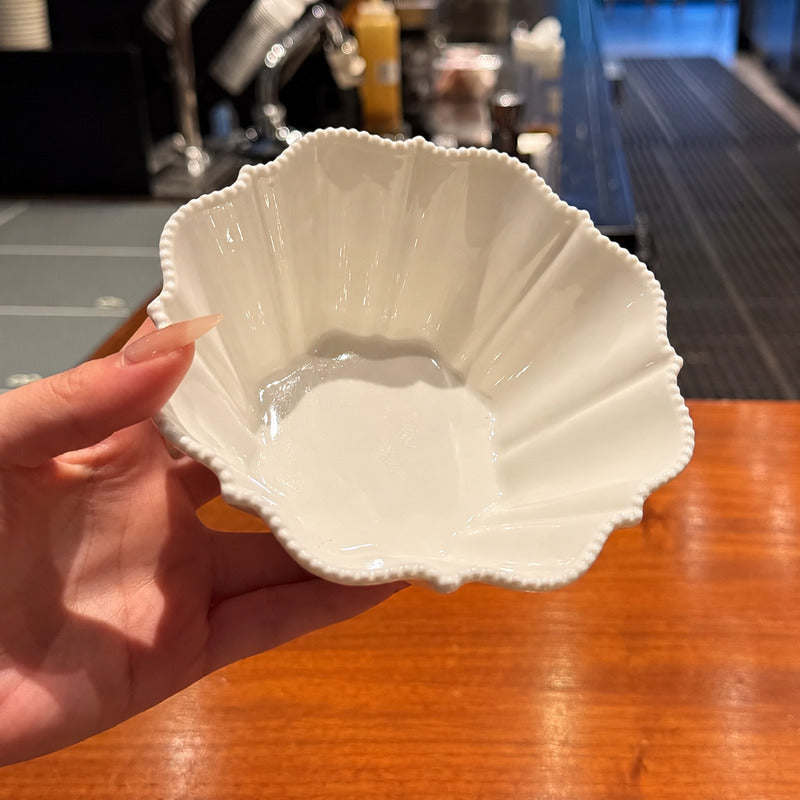A hand holding a white ceramic bowl with a scalloped rim and fluted interior, photographed over a wooden table with a blurred cafe or kitchen background.