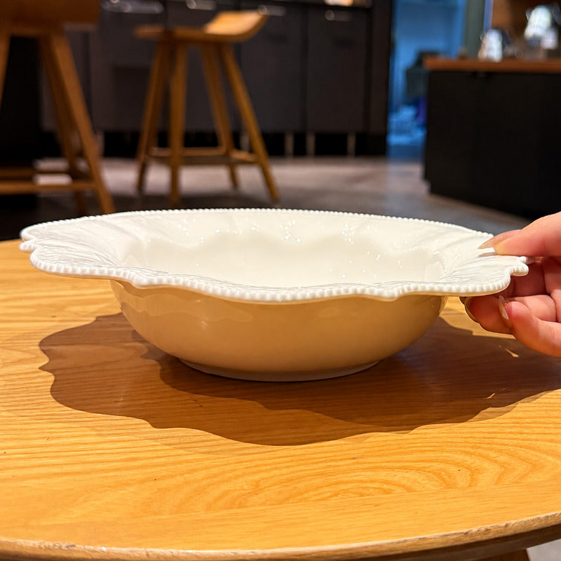 A hand holding the edge of a cream-colored ceramic bowl with a scalloped rim, set on a wooden table in a kitchen environment.