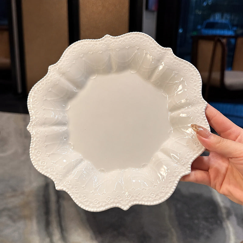 A hand holding a round white ceramic plate with a decorative scalloped edge and embossed floral pattern, set against an indoor background.