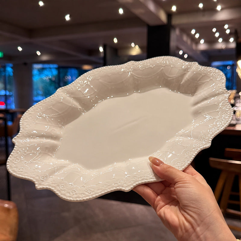 A hand holding a large, scalloped oval ceramic serving platter with a glossy white finish and embossed floral detailing along the rim, set against a blurred indoor dining background.