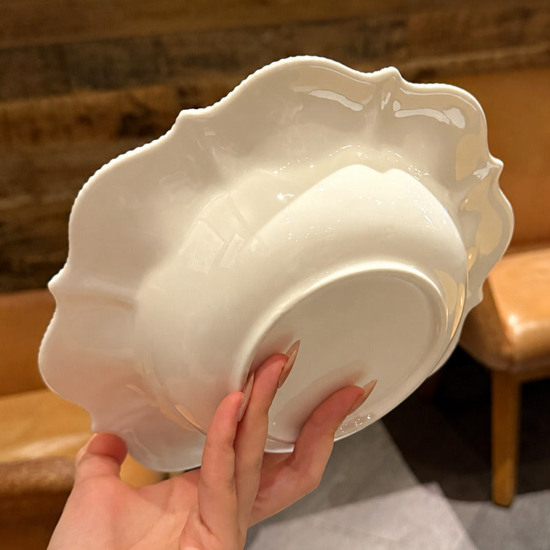 A hand holding a glossy white ceramic bowl with a decorative scalloped rim, photographed indoors against a warm brown wooden and leather background.