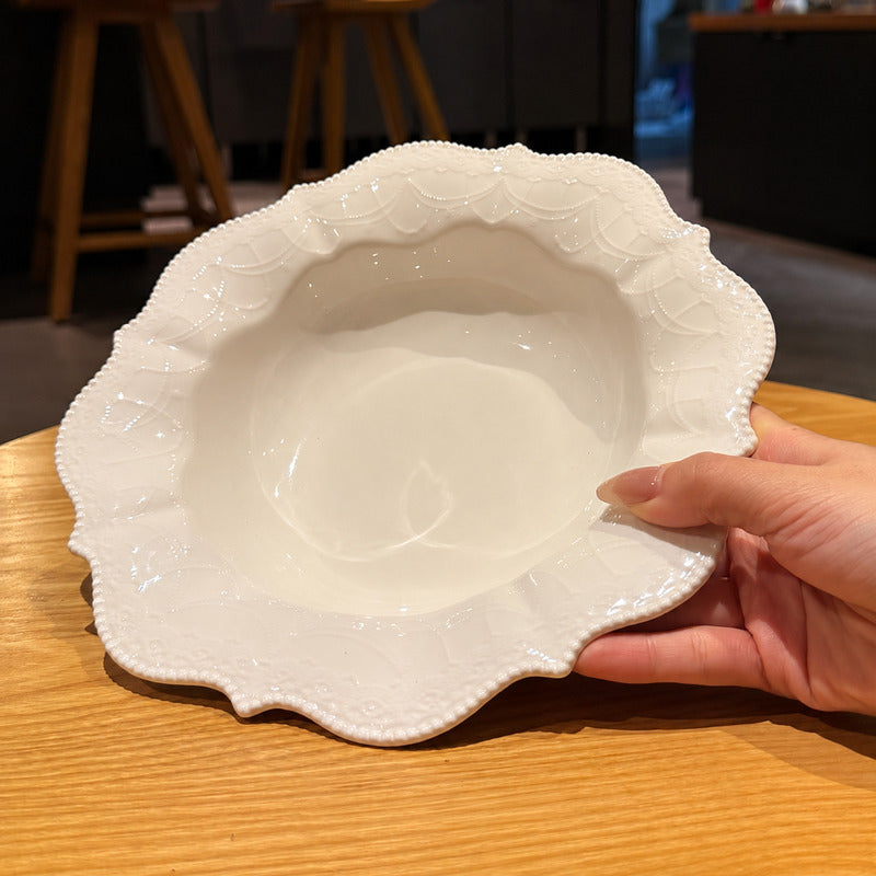 A hand holding a white ceramic bowl with a scalloped, textured rim, set on a wooden table with a blurred indoor background.