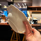 A hand holding a glossy white ceramic bowl with a decorative scalloped rim, photographed in a real indoor cafe or restaurant setting with blurred lights and wooden stools in the background.