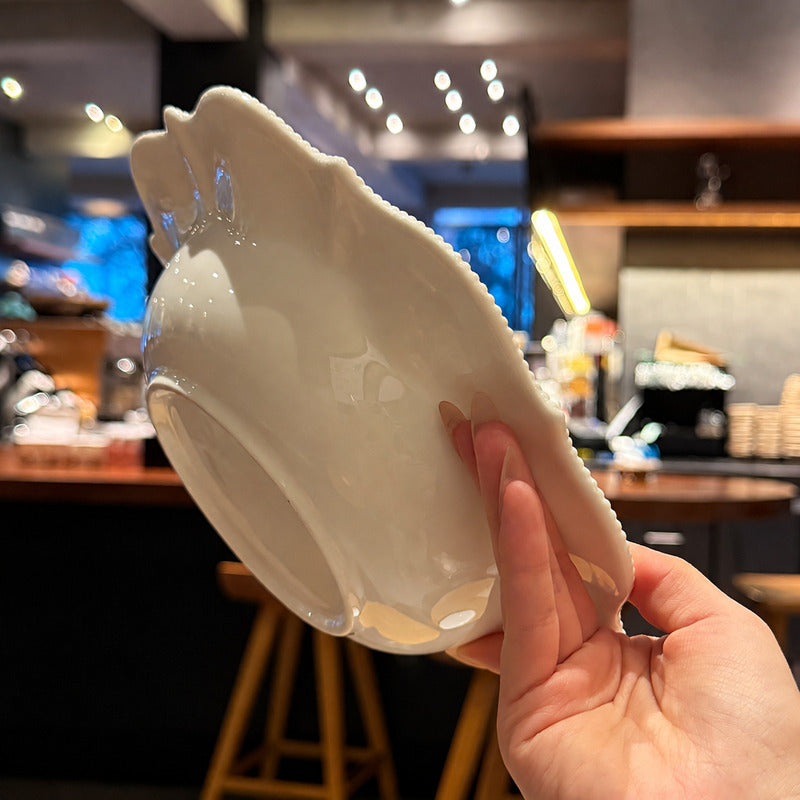 A hand holding a glossy white ceramic bowl with a decorative scalloped rim, photographed in a real indoor cafe or restaurant setting with blurred lights and wooden stools in the background.