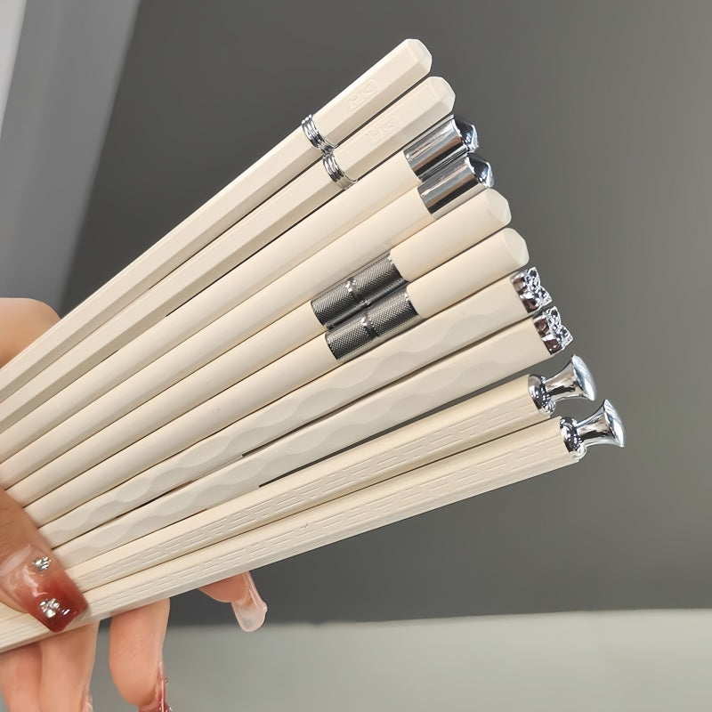 A hand holding a fan of light natural wood chopsticks with polished silver metal ferrules and tips, set against a neutral gray background.