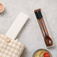 A set of wooden chopsticks and a spoon with black handles, displayed in a long rectangular white case on a light gray surface. A matching fabric pouch with a wooden button is partially visible on the left, and small bowls of food appear at the corners.