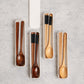 A set of wooden tableware utensils, including chopsticks, spoons, and a long-handled spoon, arranged in slim white plastic trays on a light gray textured surface.
