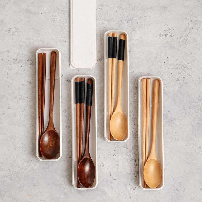 A set of wooden tableware utensils, including chopsticks, spoons, and a long-handled spoon, arranged in slim white plastic trays on a light gray textured surface.