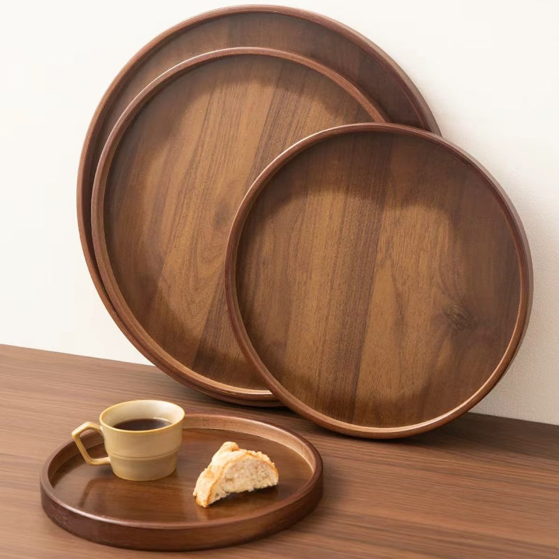 Three round wooden serving trays in varying sizes, with two stacked in the background and one in the foreground holding a cup of coffee and a piece of bread on a wooden table.