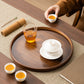 A round dark wood tea tray set on a woven mat, holding a white lidded teapot on a saucer, a clear glass cup of amber tea, and two small white teacups. A hand in a brown sleeve holds another cup at the top right.
