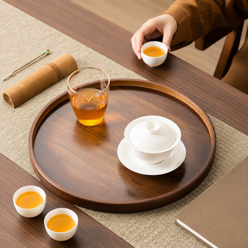 A round dark wood tea tray set on a woven mat, holding a white lidded teapot on a saucer, a clear glass cup of amber tea, and two small white teacups. A hand in a brown sleeve holds another cup at the top right.