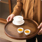 A person in a brown button-up shirt holds a round dark wood tea tray carrying a white lidded teapot and two small white cups filled with amber tea, set in a warm indoor environment.