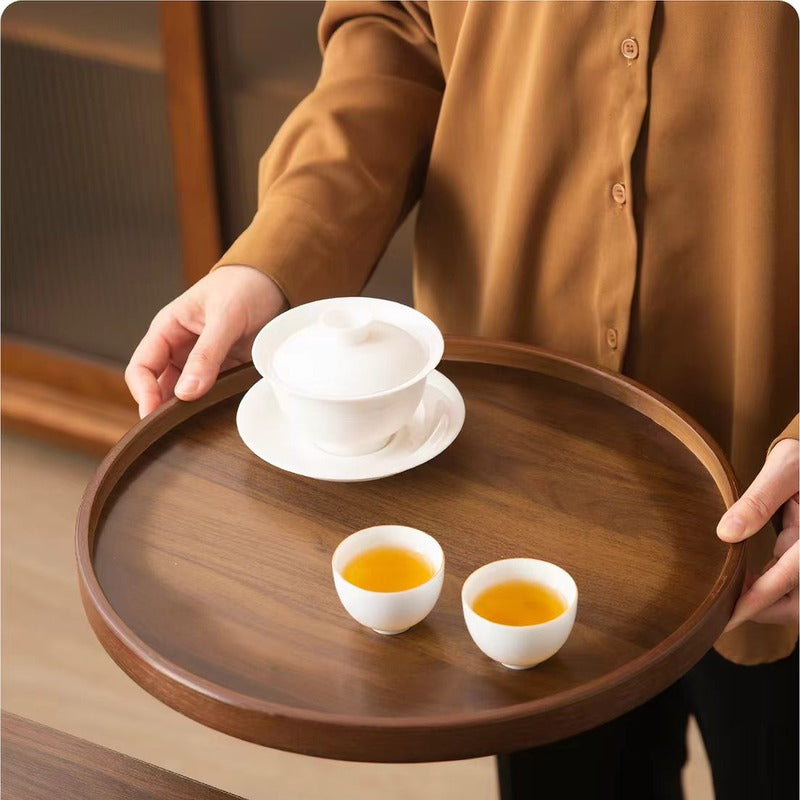 A person in a brown button-up shirt holds a round dark wood tea tray carrying a white lidded teapot and two small white cups filled with amber tea, set in a warm indoor environment.