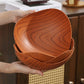 A stack of three oval wooden bowls with a rich reddish-brown grain pattern, held by two hands in a warm, rustic kitchen setting.