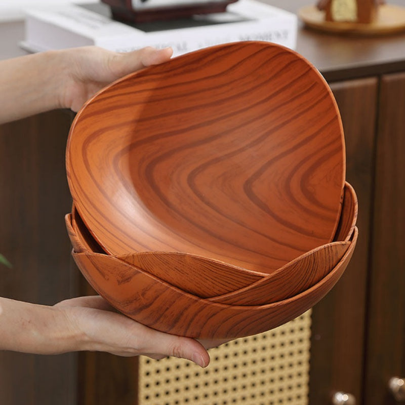 A stack of three oval wooden bowls with a rich reddish-brown grain pattern, held by two hands in a warm, rustic kitchen setting.