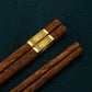 Four wooden chopsticks with dark brown grain and gold metal bands near the ends, arranged diagonally on a dark textured background.